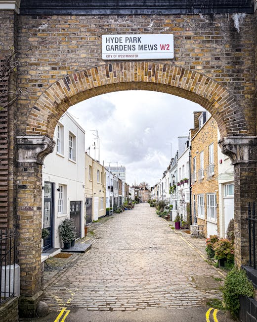 A narrow cobblestone street viewed through a brick archway with a sign reading 'Hyde Park Gardens Mews W2.' On either side of the street, there are rows of white and cream-colored terraced houses with